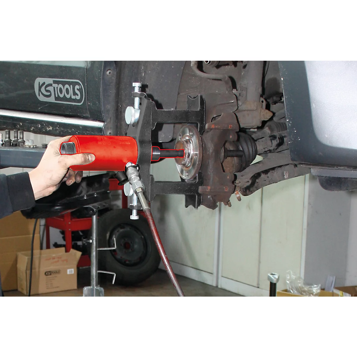 Ein Mechaniker verwendet ein rotes Werkzeug, um an einem Fahrzeug zu arbeiten. Der Fokus liegt auf der Radaufhängung und den Werkzeugen.